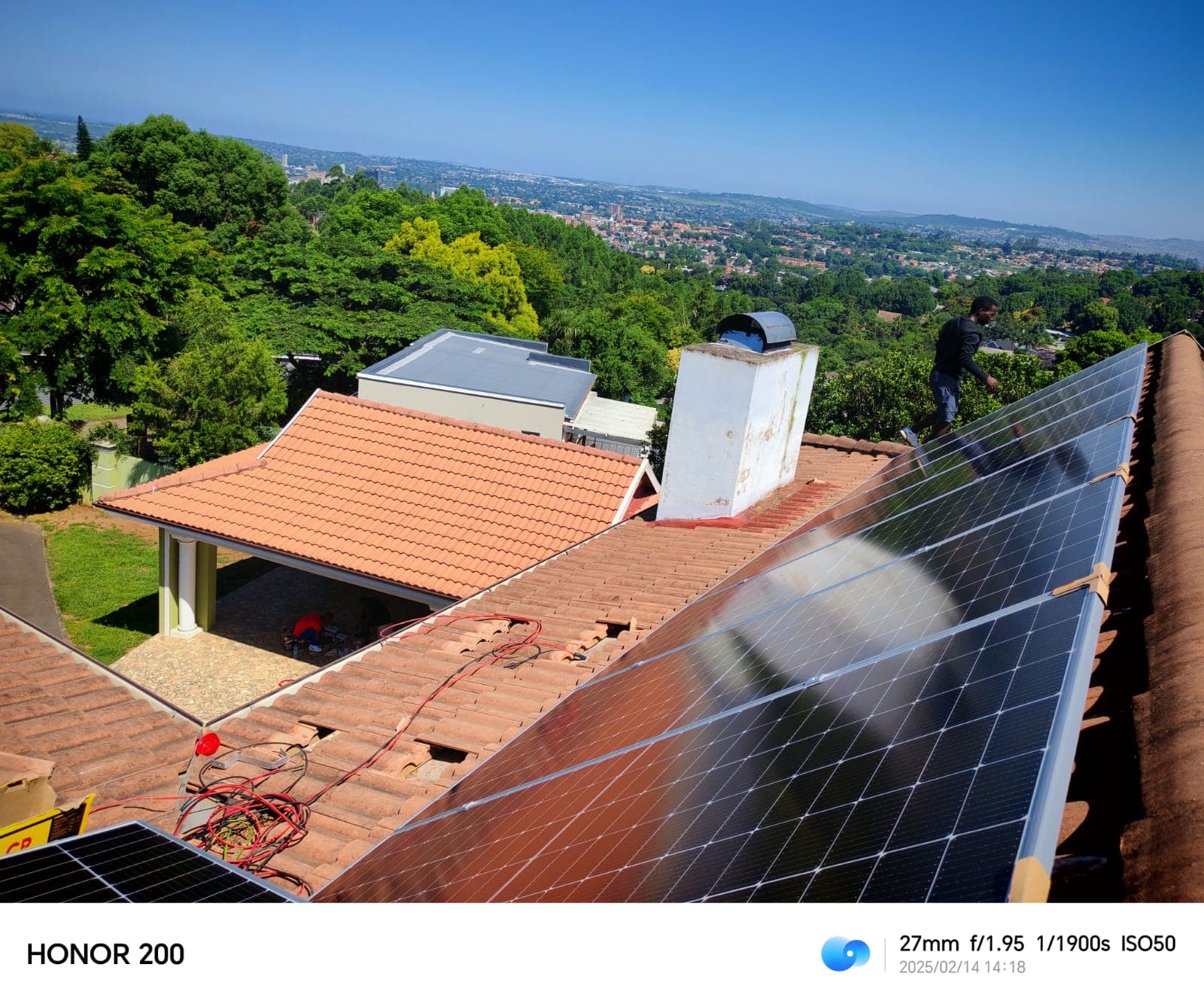 Technician installing solar panels on a red tiled roof with a scenic city background.