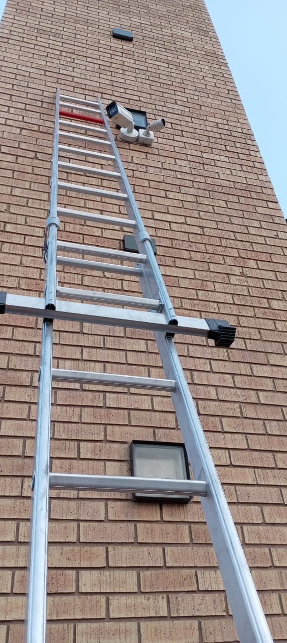 A tall silver extension ladder leans against a brick wall, reaching toward mounted security cameras.