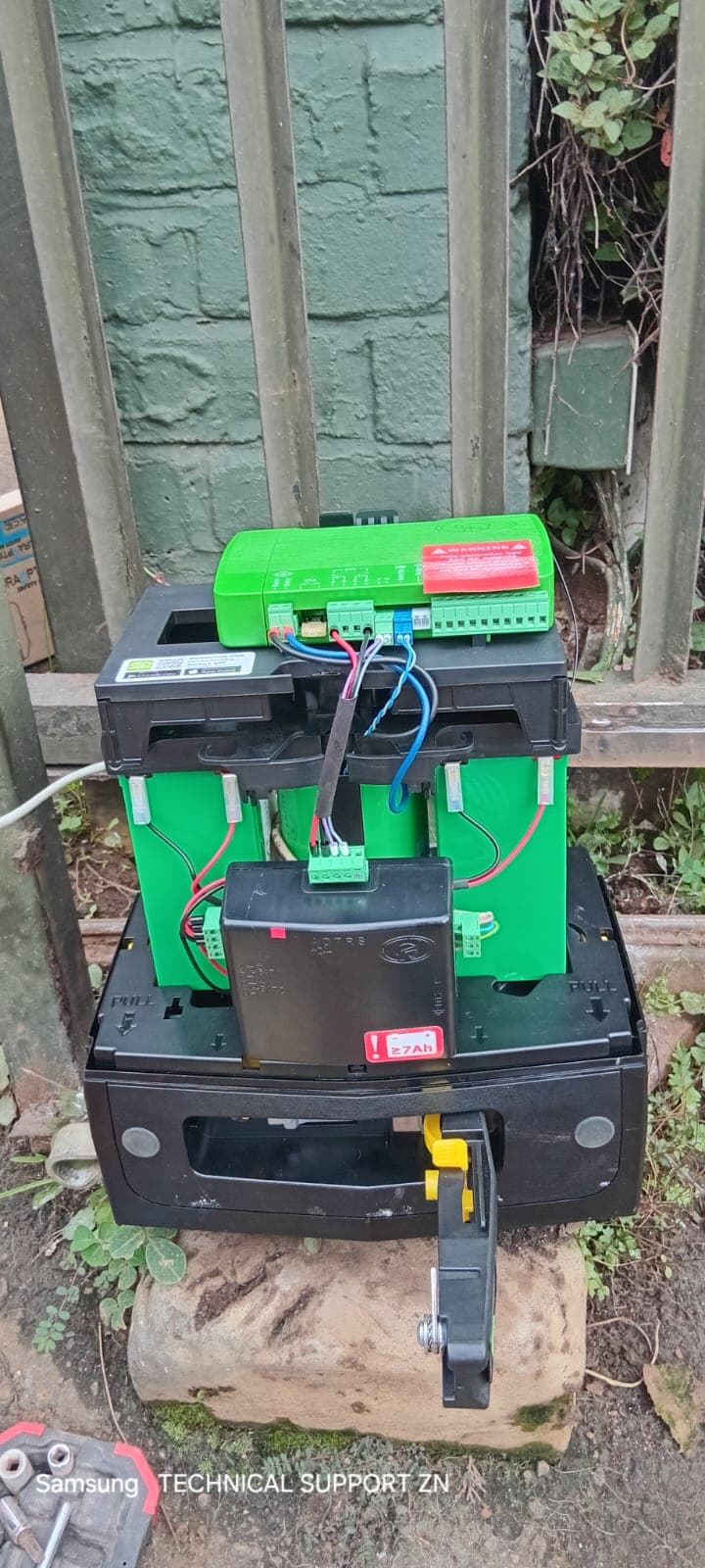 Exposed internal components of a gate motor showing green batteries and a control board outdoors.
